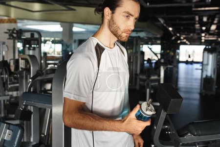 Ein athletischer Mann in aktiver Kleidung macht eine Pause und hält eine Flasche Wasser in einem Fitnessstudio, umgeben von Trainingsgeräten.