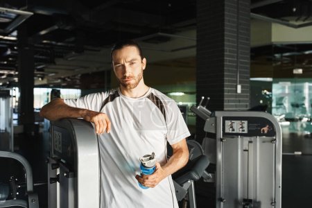Un homme athlétique en tenue active debout à côté d'une machine dans un gymnase, se préparant à sa routine d'entraînement.