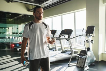 Ein athletischer Mann in aktiver Kleidung hält selbstbewusst eine Wasserflasche im Fitnessstudio.