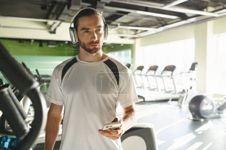 Ein athletischer Mann in aktiver Kleidung, der über Kopfhörer Musik hört und in einem lebhaften Fitnessstudio trainiert.