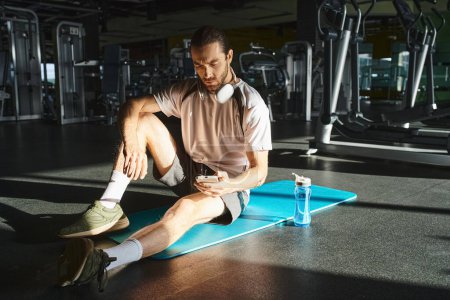 An athletic man in active wear is sitting on a blue mat in a gym.