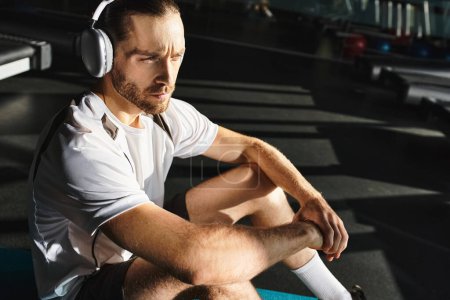 A man in active wear sits in gym, wearing headphones and lost in his music.