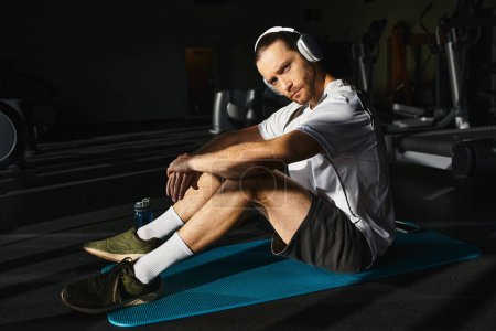 An athletic man in active wear is sitting on a blue mat, focused and calm, in the middle of a gym.