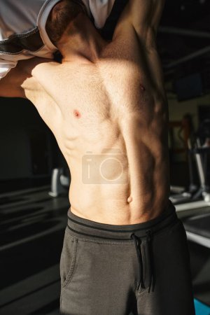 Un homme musclé sans chemise est debout dans une salle de gym, mettant en valeur son physique sculpté pendant qu'il s'entraîne.