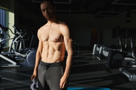A muscular man without a shirt, standing confidently in a modern dark gym.