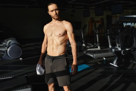 Un hombre musculoso sin camisa, de pie con confianza en un gimnasio.