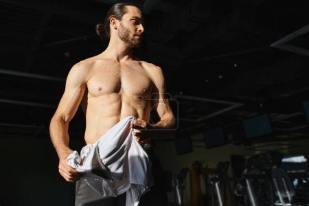 Foto de Muscular man in gym standing shirtless and holding towel in dark gym - Imagen libre de derechos