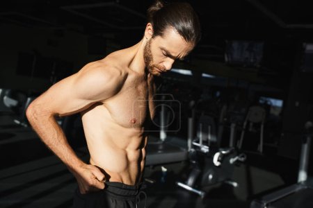 Foto de Un hombre sin camisa se para con confianza frente a una máquina de gimnasio, mostrando su físico muscular. - Imagen libre de derechos