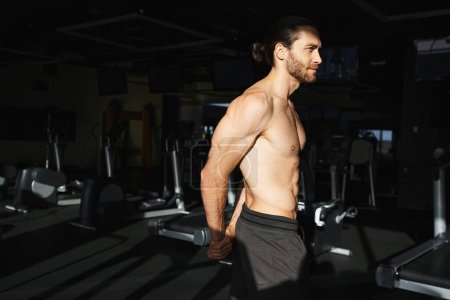 Muscular man stands confidently in gym, shirtless, showcasing his toned physique.