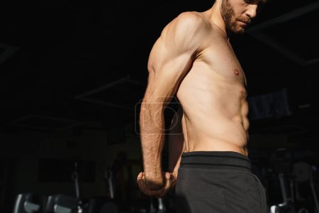 Un hombre sin camisa con músculos tonificados está de pie en un gimnasio, centrado en su rutina de ejercicios.