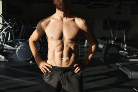 Hemdloser muskulöser Mann steht selbstbewusst im Fitnessstudio, die Hände auf den Hüften.