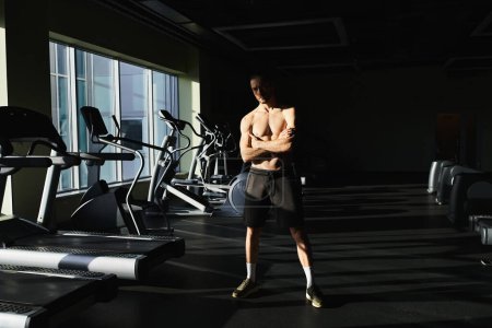 Un hombre musculoso sin camisa parado con confianza delante de una fila de cintas de correr en un gimnasio.