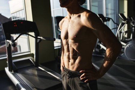 Un musculoso sin camisa se para con confianza junto a una cinta de correr en un gimnasio.