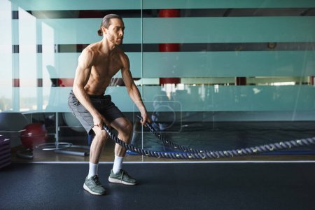 Un hombre musculoso sin camisa sostiene intensamente una cuerda de batalla en un gimnasio, mostrando su fuerza y determinación.
