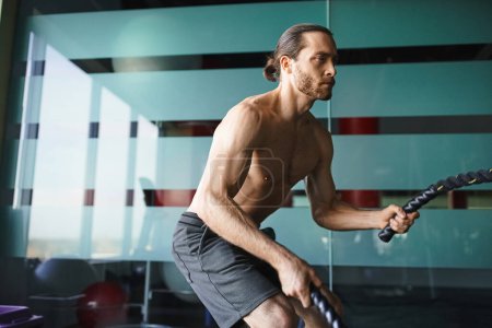 Un hombre musculoso y sin camisa sostiene con confianza una cuerda de batalla en un gimnasio.