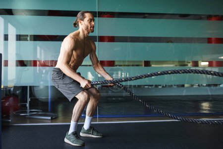 Un hombre musculoso sin camisa está agarrando y tirando de una cuerda de batalla en un gimnasio, mostrando su fuerza y determinación.