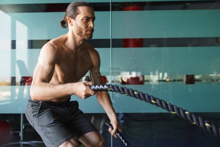 Un hombre musculoso sin camisa en un gimnasio sosteniendo una cuerda mientras ejercita su fuerza en la parte superior del cuerpo.