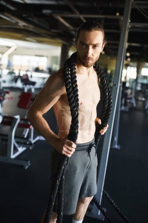 hombre sin camisa mostrando físico muscular mientras se agarra una cuerda en el gimnasio.