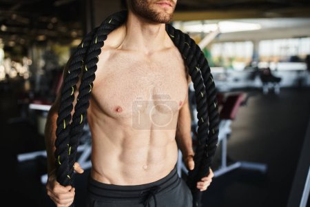 A muscular man without a shirt fiercely holds a rope, showcasing his strength and determination during a workout session in the gym.