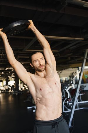 Shirtless man displaying power, lifting weight plate in gym.
