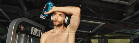 Muskelprotz ohne Hemd hält eine Flasche Wasser in der Hand und trägt Kopfhörer für seine Trainingseinheit.