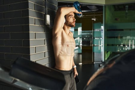 Muscular shirtless man takes a refreshing break, holding a bottle of water after an intense workout in the gym.