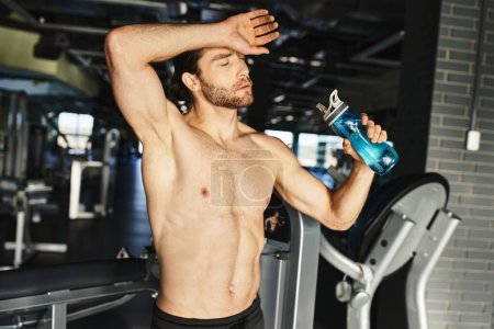 Strong shirtless man taking a break, holding a bottle of water in a gym setting.