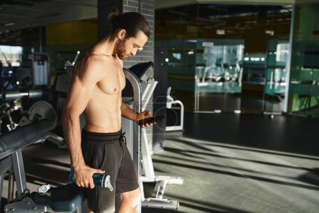 Hombre sin camisa en el gimnasio, intensamente enfocado en la pantalla del teléfono celular mientras hace ejercicio.