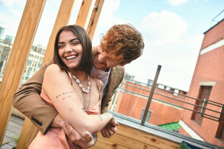 Téléchargez les photos : Un homme enroule ses bras autour d'une femme dans un câlin serré sur un toit surplombant la ville, exprimant amour et connexion - en image libre de droit