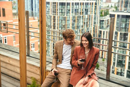 Un couple partage un moment de rire et de vin rouge, encadré par une vue sur le paysage urbain sur un balcon en bois