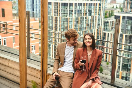 Téléchargez les photos : Un couple souriant se détend avec un verre de vin, embrassé par la chaleur du soleil couchant et le paysage urbain - en image libre de droit