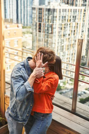 A man and a woman share a tender kiss on a balcony overlooking a cityscape, their bodies pressed together in an intimate embrace