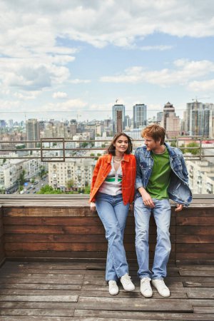 Un hombre y una mujer se paran triunfantes sobre un rascacielos, mirando a la ciudad de abajo con una mezcla de asombro y orgullo