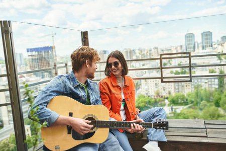 Ein Mann mit einer Gitarre singt einer lächelnden Frau auf einem Dach über der städtischen Skyline zu.