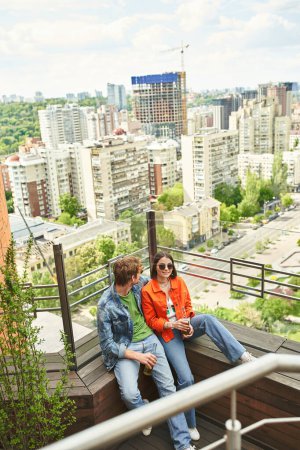 Téléchargez les photos : Un couple de personnes s'assoient paisiblement sur le bord d'un haut bâtiment, profitant de la vue sur les toits de la ville au crépuscule - en image libre de droit