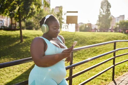 Una mujer afroamericana, abrazando su positividad corporal, usando auriculares mira atentamente su teléfono celular mientras disfruta de la música al aire libre.
