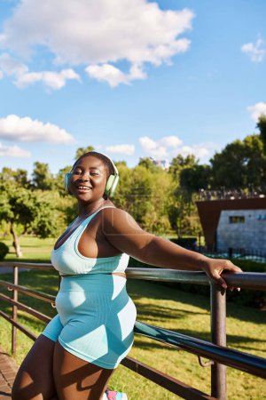 Eine Afroamerikanerin mit Kopfhörern lehnt in einem blau-weißen Outfit an einer Schiene und verkörpert Körperpositivität.