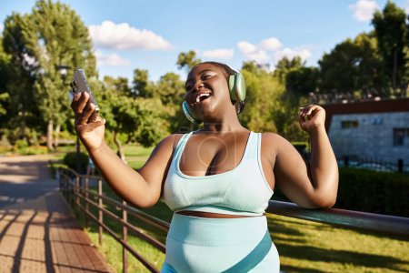 An African American woman, body positive and curvy, wearing headphones while holding a cell phone outdoors.