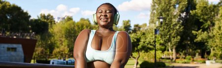 Una mujer afroamericana con curvas en una parte superior de pie con confianza en un parque, escuchando música a través de auriculares y abrazando su positividad corporal.
