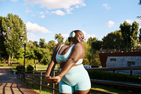 An African American woman with a curvy body, wearing a blue sports bra and shorts, listening to music outdoors.