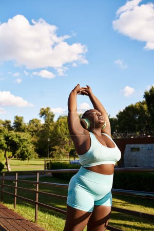 An African American woman in a blue top and shorts energetically stretches her arms outdoors, embodying body positivity.