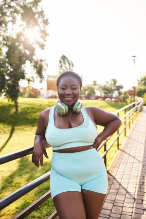 Photo for An African American woman with headphones, wearing a blue top and shorts, confidently poses for a picture outdoors. - Royalty Free Image