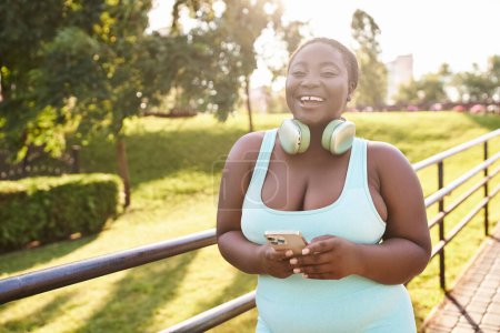 A confident African American woman, wearing headphones, enjoys music while holding a cell phone outdoors.