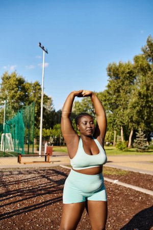 A curvy African American woman in a blue sports bra top stretching her arms while exercising outdoors.
