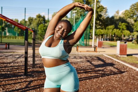 An African American woman with curvy body, in blue sports bra, stretching her arms outdoors.