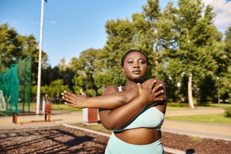 Curvy African American woman in sportswear standing gracefully in a park, stretching her arms outdoors.