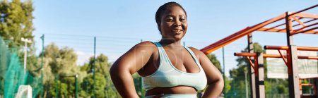 An African American woman in sportswear stands confidently in front of a playground, embracing her body positivity and strength.