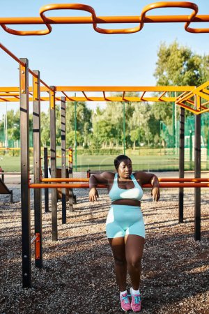Foto de Una mujer afroamericana en ropa deportiva azul se encuentra con confianza en un patio de recreo, encarnando la positividad corporal y la libertad. - Imagen libre de derechos