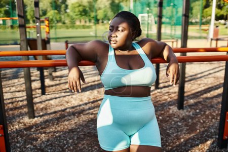 Une femme afro-américaine en tenue de sport se tient joyeusement devant une aire de jeux, incarnant la positivité et l'autonomisation du corps.