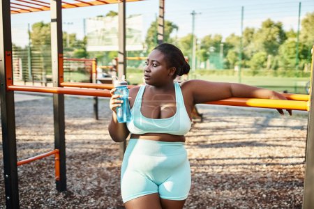 An African American woman in sportswear holds a water bottle while enjoying a moment of hydration in a park.